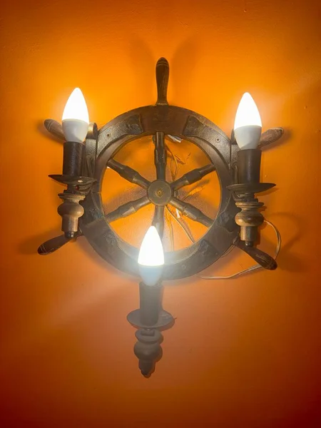 Turuncu bir duvara monte edilmiş dekoratif gemi tekerleği lambası. Tesisatta üç tane patlamış ampul var..