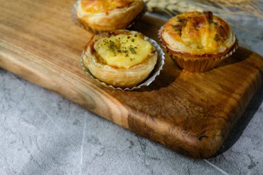 Üç tane ev yapımı, altın hamurlu ve kremalı tart, ahşap bir tahtaya, temiz desenli arka planda yerleştirilmiş. Fırın promosyonları ve tatlı menüleri için mükemmel.