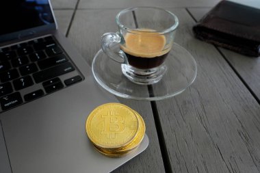 Altın Bitcoin, bir espresso bardağının yanına, tahta bir masadaki dizüstü bilgisayara yerleştirilmiş..