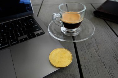 Tabağın üzerinde bir fincan espresso. Tahta masadaki dizüstü bilgisayardaki altın bir Bitcoin 'in yanında..