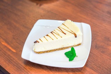 Zarif bir çikolata yağmuru ve taze bir nane yaprağı ile süslenmiş, el değmemiş beyaz bir tabakta sunulmuş nefis bir dilim klasik cheesecake..