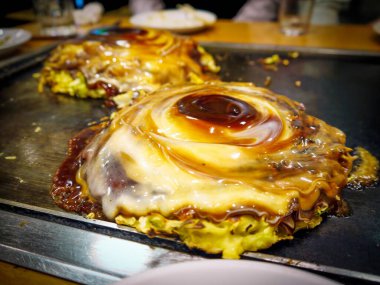 İki lezzetli Japon leziz gözlemesi, Okonomiyaki, sıcak teppan ızgarasında cızırdıyor, üzerinde bol sos ve kremalı mayonez kıvrımları var..