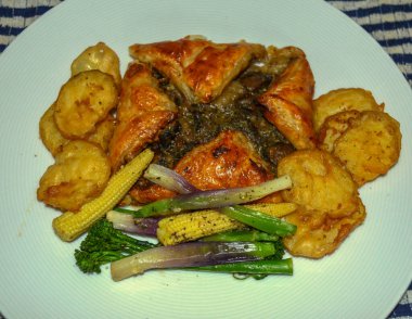 Lezzetli bir mantar dolu hamur işi. Etrafı altın kıtır patates dilimleri, renkli kızarmış sebzeler ve taze taze taze soğanlarla çevrili. Mavi-beyaz çizgili arka planda..