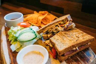 Kabuklu, katmanlı et, peynir ve sebzeli bir panini, çıtır salatalık, domates ve ahşap bir tahtanın üzerinde iki kremalı sosla servis ediliyor..
