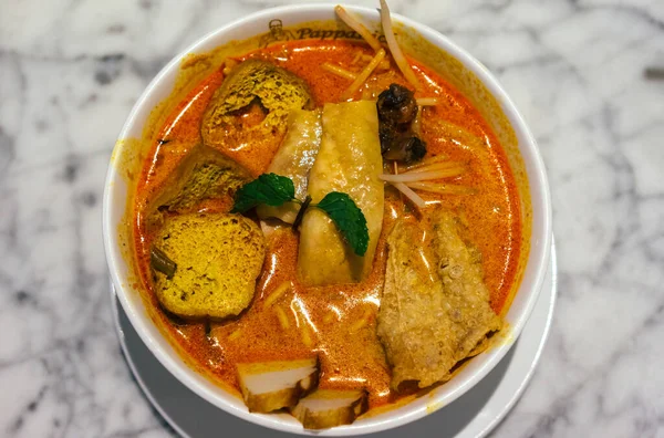 Beyaz bir kâse içinde PappaRich logosu olan, parlak mermer bir tezgahın üzerine yerleştirilmiş beyaz bir kase Malezya körili laksa eriştesi..