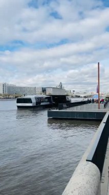 riverside ferry dock under cloudy sky, modern passenger boat moored at concrete railing, calm water reflections, distant city skyline, muted winter light, slow commuter atmosphere