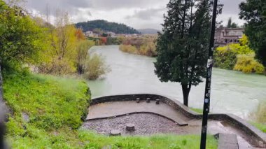 park terrace overlooking river bend with mature tree and curved stone promenade, paved viewpoint with railing, grassy slope