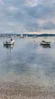 Çakıl taşı plajı, demirli sandal ve berrak su, sığ kıyı şeridi taşları ortaya çıkarıyor, bulutlu gökyüzünün altında sakin bir koy.,