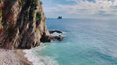 Dramatik uçurum koyuna karşı turkuaz su, bitki örtüsünün altında çakıl taşı plajı ufukta kayalık yüz, uzak ada,