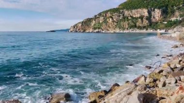 Kayalık kıyı şeridi, dalgalı turkuaz dalgalar, kayaların üzerinde patlayan enerjik dalgalar, dokulu ön plan taşları ve uzak yeşil