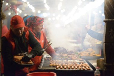 12 / 21 / 2024 Craiova, Romanya: Buhar yükselirken ve müşteriler mevsimsel şenlikler sırasında toplanırken, gıda satıcıları dumanlı bir Noel pazarında et ızgara ediyorlar