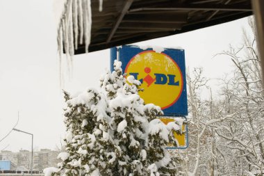 16 Mart 2024 'te Bulgaristan' ın Rusçuk kenti: Karla kaplı bir Lidl mağaza tabelası, kış ağaçları ve sarkıtların yanında duruyor, yoğun kar yağışı süpermarket alanı etrafındaki manzarayı şekillendiriyor.