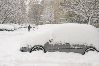 Ağaçlar, binalar ve taze karla kaplı park edilmiş araçlarla yoğun kış koşullarında yürüyen bir insan, yoğun bir cadde boyunca derin bir karın altında gömülü olarak bulunuyor..
