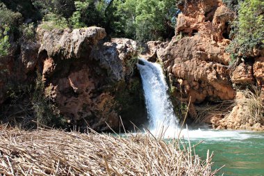 Tavira, Algarve, Portekiz 'deki kırmızı kayalık kayalıklar ve yeşil bitki örtüsüyle çevrili Pego do Inferno şelalesinin yakın görüntüsü. Güneşli bir yaz gününde.