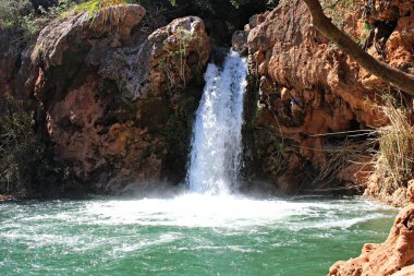 Tavira, Algarve, Portekiz 'deki yeşil bitki örtüsüyle çevrili kırmızı kayalıklara tırmanan kişi ile Pego do Inferno Şelalesi manzarası. Aydınlık bir yaz gününde.