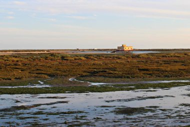 Ria Formosa Doğal Parkı, Fuseta, Algarve, Portekiz 'deki gelgit sulak alanları ve sembolik sarı evlerin günbatımı manzarası.