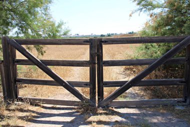Ahşap kapı, kırsal alanda, parlak güneş ışığı altında kuru çimenler ve yeşil çalılarla çevrili bir toprak yolu kapatıyor..