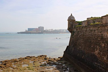 İspanya 'nın Cadiz kentinde Castillo de San Sebastian' ın tarihi kıyı kalesi, antik taş duvarları ve kayalık kıyı şeridinin üzerinde alçak gelgitle yükselen gözlem kulesi. Körfezin sakin suları ve uzak modern şehir bulutlu bir gökyüzünün altında ufuk çizgisi.