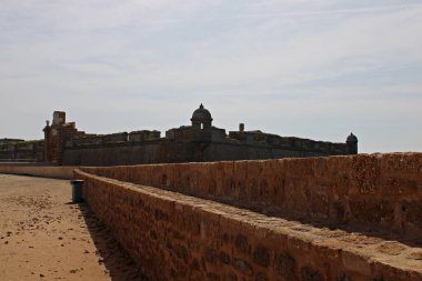 İspanya 'nın Cadiz kentinde Castillo de San Sebastian' ın antik taş duvarları ve savunma kulesi sisli bir gökyüzünün altındaki sahil geçidinden bakıldığında, tarihi yapılar, mimari ve kıyı bölgelerini alçak gelgitte sergiliyor.