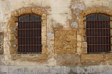 İspanya 'nın Cadiz kentindeki Castillo de San Sebastian' da iki eski parmaklıklı pencereli yıpranmış taş cephe. Yaşlanmış dokular, soyulmuş alçı, aşınmış kumtaşı blokları ve paslanmış demir çubuklar tarihi bir kıyı atmosferi yaratır.