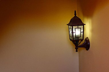 Aydınlatılmış eski tarz duvar feneri, sıcak ortam ışığını bir köşedeki iç mekâna yayıyor, rahat gölgeler ve sakin bir atmosfer yaratıyor, yumuşak sarı duvarlar ve ev girişleri, verandalar ve koridorlar için uygun klasik dekoratif bir tasarım.