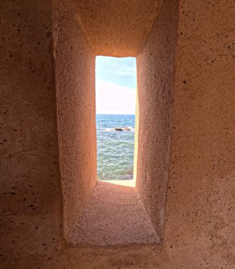 İspanya 'nın Cadiz kentindeki Castillo de San Sebastian antik gözlem kulesinin içinden görülen mavi okyanus dalgaları ve kayalık kıyı şeridi üzerindeki parlak güneş ışığını çerçeveleyen tarihi bir kıyı kalesinin içindeki dar bir taş gözcü yuvası..