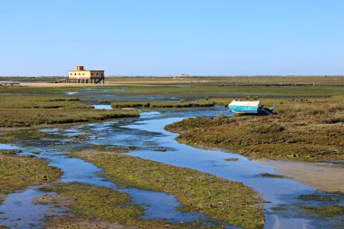 Fuseta, Algarve, Portekiz 'deki Ria Formosa tuzlu bataklıklarının manzarası, sığ gelgit kanalları, yeşil deniz yosunu, kıyı bitkileri, terk edilmiş küçük bir balıkçı teknesi ve durgun, güneşli bir günde açık mavi bir gökyüzünün altında durgun bir deniz binası.