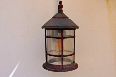 Eski kırsal sokak lambası desenli bir duvara monte edilmiş, yıpranmış metal, eski cam paneller ve sıcak bir filaman ampulü, geleneksel Avrupa kentsel aydınlatmasının cazibesini ve tarihi sokakların ebedi karakterini yansıtıyor.