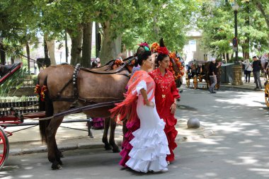 Sevilla Nisan Fuarı 'na giderken süslü at arabasının yanında poz veren geleneksel Sevilya elbiseli Flamenko kadınlar, ağaçlar ve şenlikli atmosferle çevrili. Endülüs kültürü, moda ve Güney İspanya 'da canlı bahar kutlaması