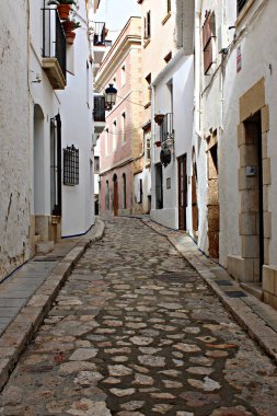 Sitges 'de kaldırımı olan dar bir ortaçağ sokağı, geleneksel İspanyol eski kasaba mimarisi, balkonlu beyaz evler, tarihi Avrupa sokağı, güney Avrupa seyahat merkezi, büyüleyici köy atmosferi, kültürel miras.