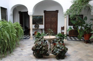 Geleneksel Endülüs bahçesinde mermer çeşme, kemer ve sütunlar, yemyeşil bitkiler ve dama tahtası tabanı, klasik İspanyol iç avlu mimarisi, zarif tarihi ev tasarımı, simetri, sükunet ve Akdeniz yaşam tarzı kavramı