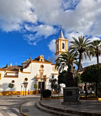 Iglesia de Santa Maria Magdalena çan kulesi, palmiye ağaçları ve Plaza de los Jardines 'deki anıt, Endülüs, İspanya, tarihi beyaz ve koyu renkli kilise dramatik mavi gökyüzü altında bulutlar, kentsel kültür simgesi