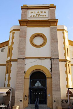 Santa Pola, Alicante, İspanya 'daki tarihi Plaza de Abastos pazar binasının önünde, bir kıyı kasabasındaki yerel ticaret, kültür ve kentsel mirası temsil eden geleneksel Akdeniz mimarisi, simetri ve sıcak renkler yer alıyor.