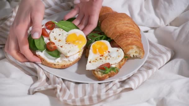 Valentijnsdag concept. Vrouw die ontbijt op bed eet op Valentijnsdag. Broodjes met croissant, bagel, roomkaas en gebakken eieren hart op wit bed. — Stockvideo