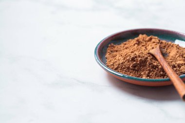 Çiğ organik kakao tozu, tahta kaşıklı, beyaz mermer kaplamalı bir çay tabağı. Süper yemek konsepti.