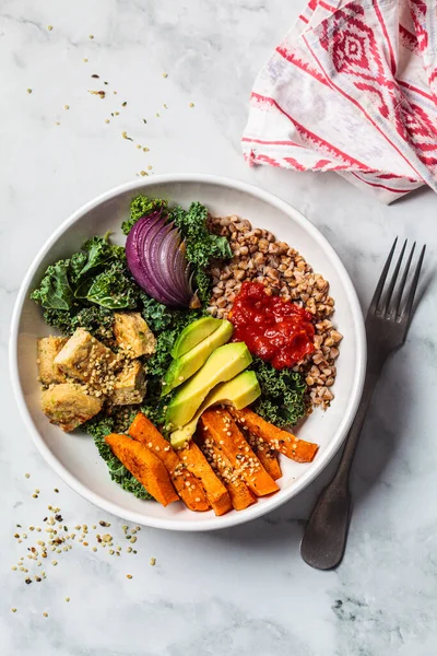 Baked vegetables, avocado, tofu and buckwheat buddha bowl. Vegan lunch salad with kale, baked sweet potato, tofu, buckwheat and avocado in a white bowl. Vegan concept.