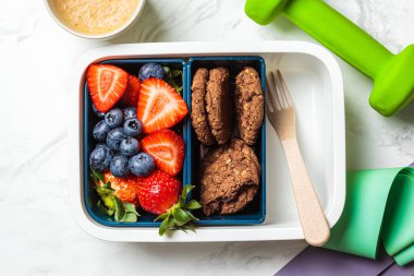 Spor aletleri ve beyaz mermer arka planda sağlıklı beslenme çantası. Paket servis için içinde meyve ve kurabiye var..