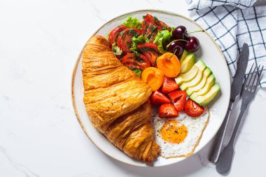 Breakfast plate with croissant, avocado, fried egg, salad and fruit, top view.