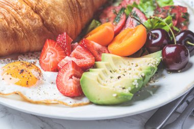 Breakfast plate with croissant, avocado, fried egg, salad and fruit, close-up.