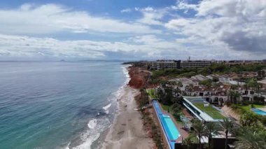 Dehesa de Campoamor 'un turkuaz deniz dalgaları, kumlu plaj, kızıl kıyı uçurumları, modern deniz manzaralı binalar ve palmiye kaplı sokaklar manzaralı Akdeniz kıyısı manzarası