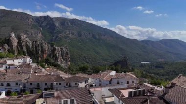 Mavi gökyüzü ve bulutlarla dolu güneşli bir günde yeşil dağlar ve kayalık kayalıklarla çevrili geleneksel beyaz bir köyün manzarası..