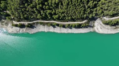 Turkuaz göl suyunun tepeden aşağı hava aracı görüntüsü kayalık kıyı şeridi, orman ağaçları ve kıyı boyunca uzanan toprak yol doğal doğa manzarası.
