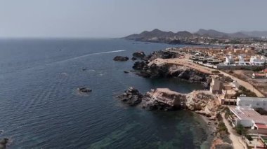 Güneşli bir günde sahil binaları, berrak deniz suyu, uçurumlar ve sahil kasabası yakınlarındaki tekne yolu ile kayalık kıyı şeridini gösteren insansız hava aracı görüntüsü.