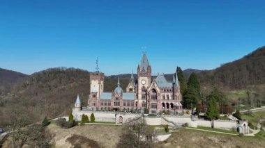 Süslü kuleleri ve taş duvarları olan tarihi şato berrak mavi gökyüzünün altında orman tepeleriyle çevrili peri masalı ve miras atmosferi yaratıyor.