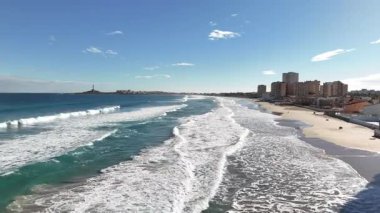 Güneşli bir günde, deniz kenarı ve sahil binaları, mavi gökyüzü ve sakin atmosferiyle kumlu kumsala yuvarlanan okyanus dalgalarının insansız hava görüntüsü..