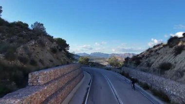 Güneşli bir günde İspanya 'nın kırsal kesimindeki kayalık tepeler ve uzak dağlar arasında dönen dağ yolunda bisikletçinin insansız hava görüntüsü..