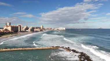 Okyanus dalgaları, apartman binaları ve taştan dalgalı sular, deniz kenarındaki güneşli deniz manzaralı kıyı kenti manzaralı İHA panoraması.