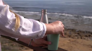 Kadının sıcak kahveyi metal termostan bardağa doldurup deniz kenarında dinlendirici bir plaj sabahı hayat tarzı ve açık hava konsepti.