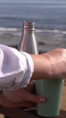 Paslanmaz çelik şişeden, deniz kıyısında sürdürülebilir yaşam tarzı yolculuğu ve hidrasyon kavramına yakın, yeniden kullanılabilir eko bardağa el yapımı içeceği dök.