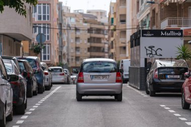 Rear view of small car driving along residential urban street lined with parked vehicles and apartment buildings in European town.